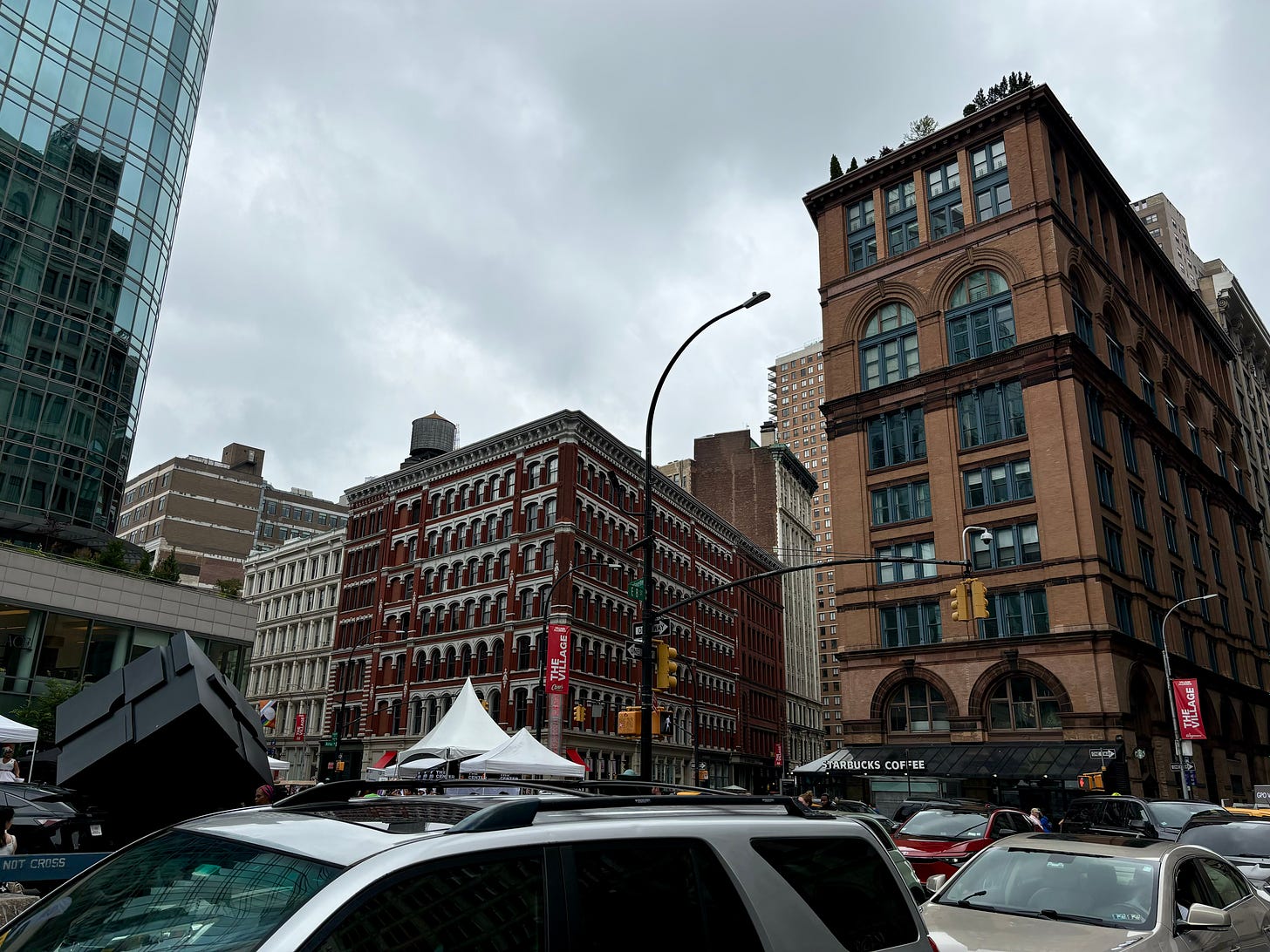 A large brown building with ornate windows and a Starbucks on the ground floor. It is across the street from a red building with a roof tank and to the left of the frame is the Astor Place cube.