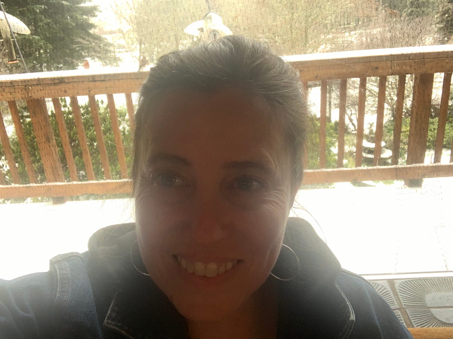 A woman smiles. Behind her is an ice-covered porch and you can just make out birds and a feeder.