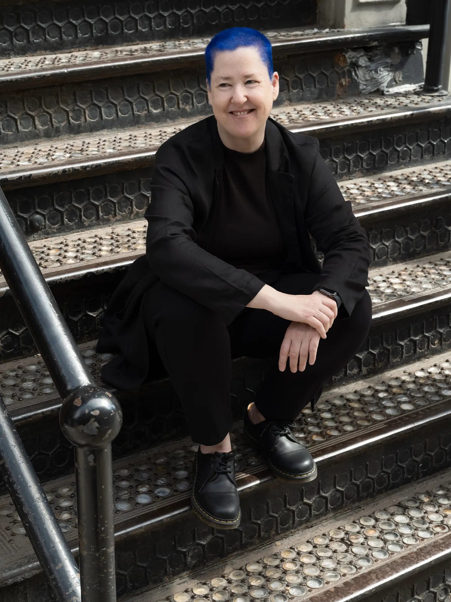Photograph of Sal Randolph, a white woman with very short, very blue hair. She is sitting on old cast-iron steps in Soho, New York, and smiling.