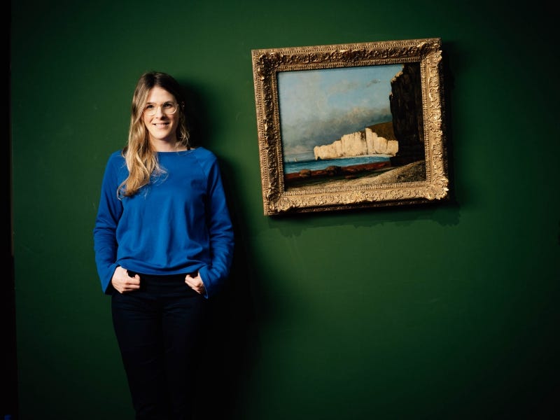 Woman standing in front of slightly tilted old painting of clifftop scene, in gilt frame, on green wall.