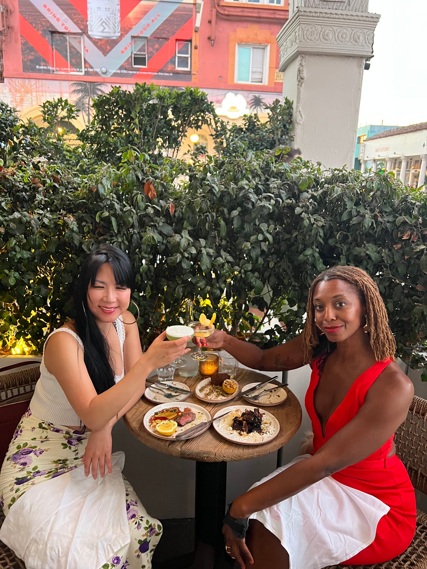 Two people, Korean and Black, in white and vermillion dresses respectively, toasting over cocktails on a patio in Venice Beach.