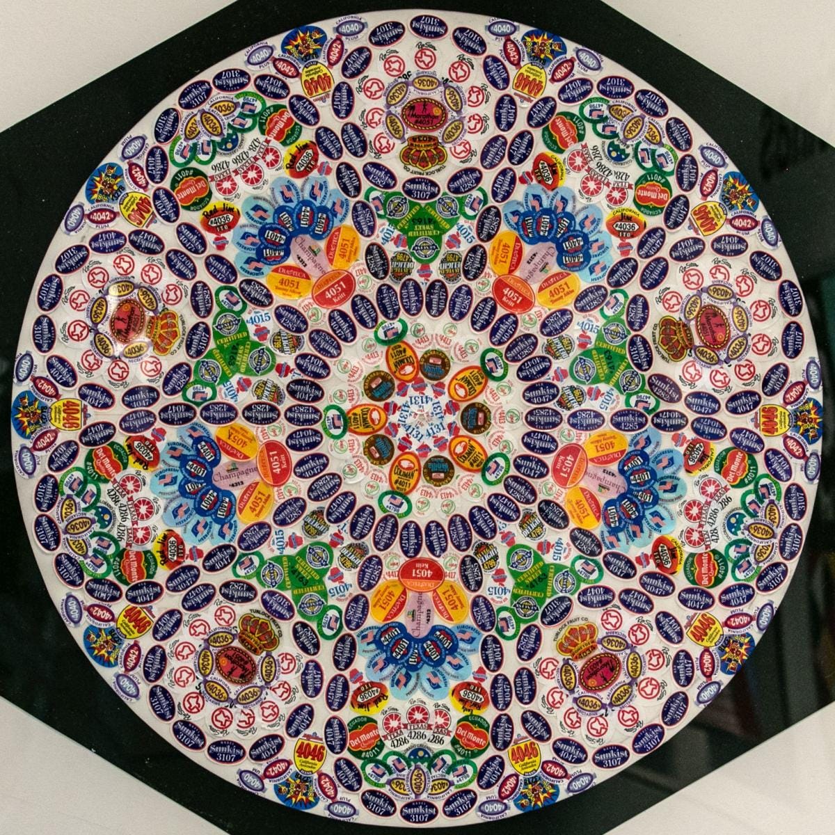 A circular, mandala-like collage made from fruit stickers that resembles a church window.