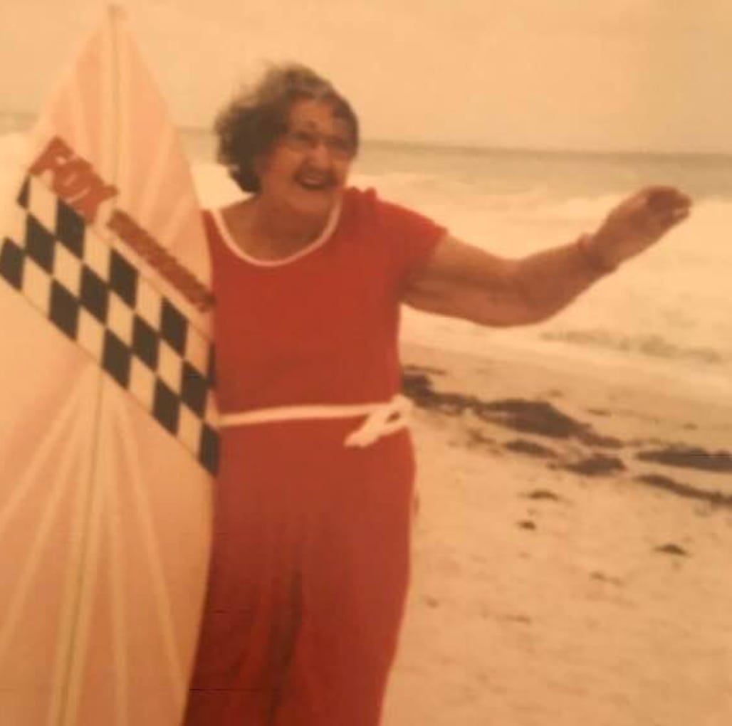 Grandma in red jumper holding a surfboard on the beach.
