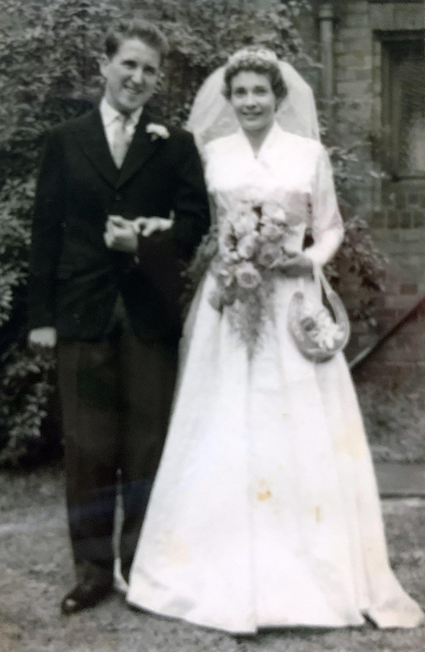 A photo of my grandparents smiling on their wedding day. My grandad in a black suit with a white flower in his button hole. My gradnma in white holding a bunch of flowers and a veil in her hair. 