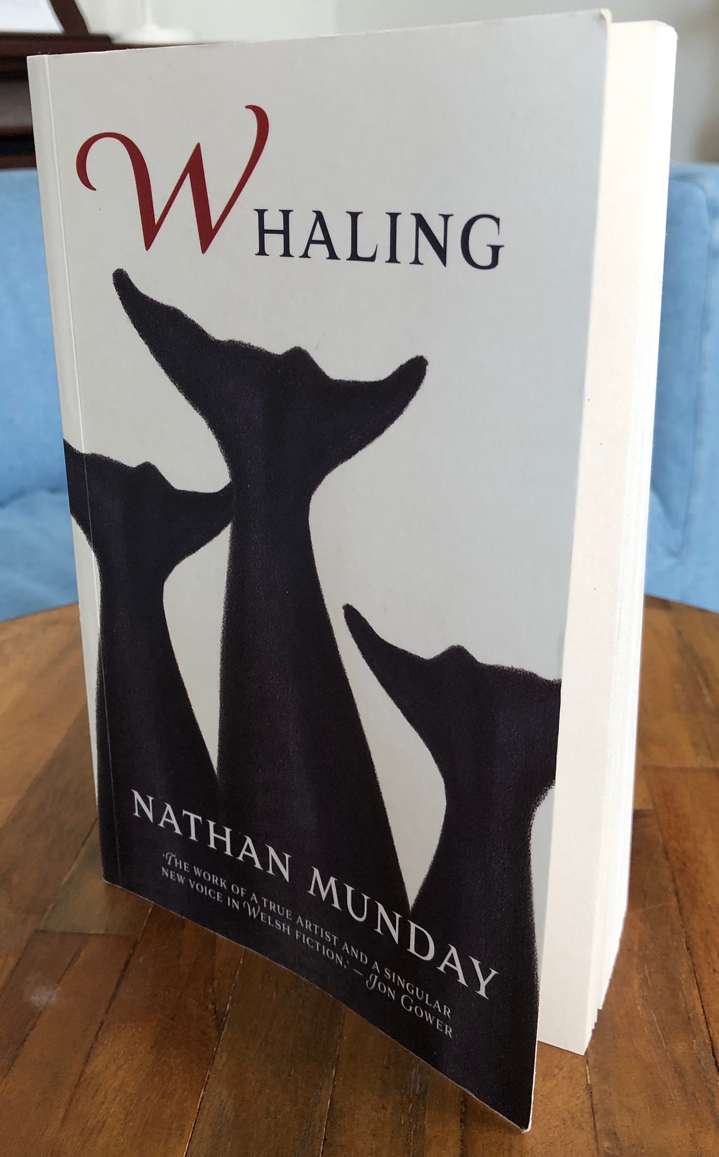 A book, Nathan Munday’s Whaling, stands on a round wooden table in front of a couch.