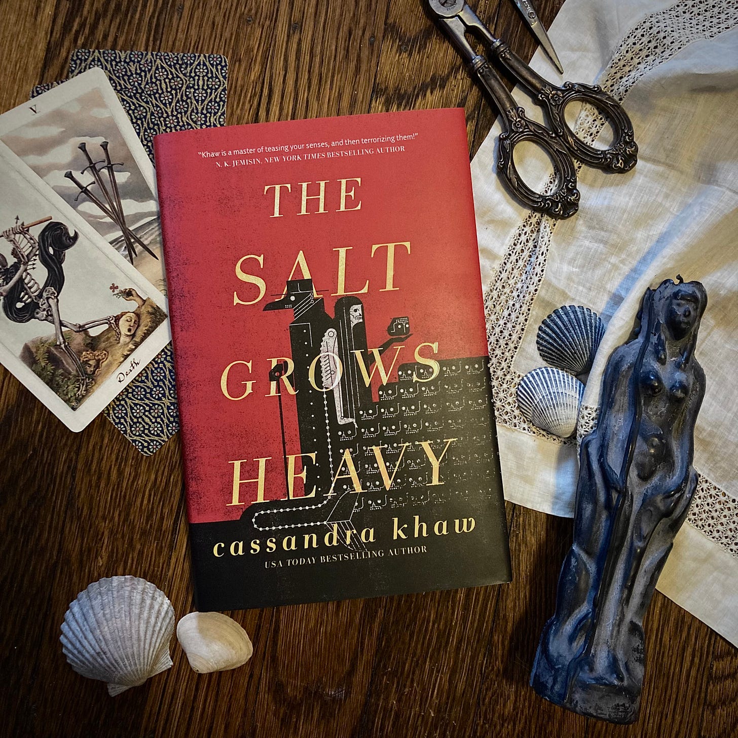 a copy of The Salt Grows Heavy in a flat lay on brown hardwood floors featuring tarot cards from the Pagan Otherworlds deck, seashells, a black candle in the shape of a woman and vintage silver scissors with an ornate handle