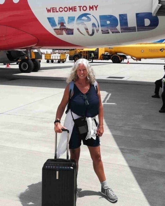 a woman with wavy grey hair standing with a roller suitcase in front of an airplane and smiling