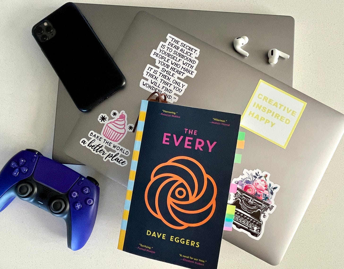 THE EVERY by Dave Eggers on top of a stack of laptops, with a phone, Airpods, and video game controller surrounding the book.