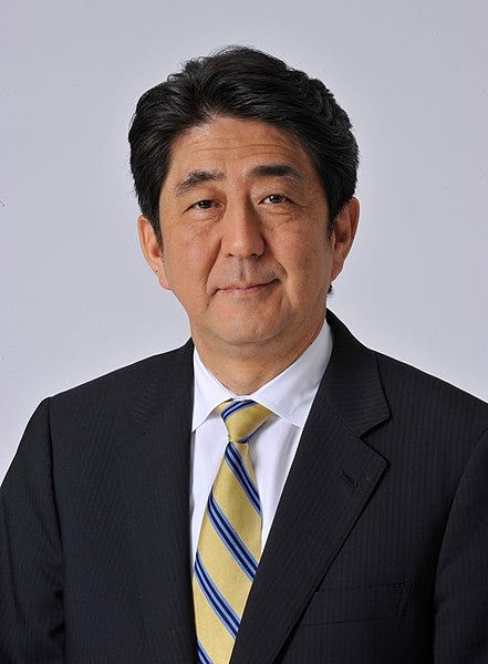 An older man with a full head of jet black hair, in a black suit with a yellow and blue striped tie stares wearily into the camera, evincing a politician's smile but his eyes remain distant. Clearly an official portrait.