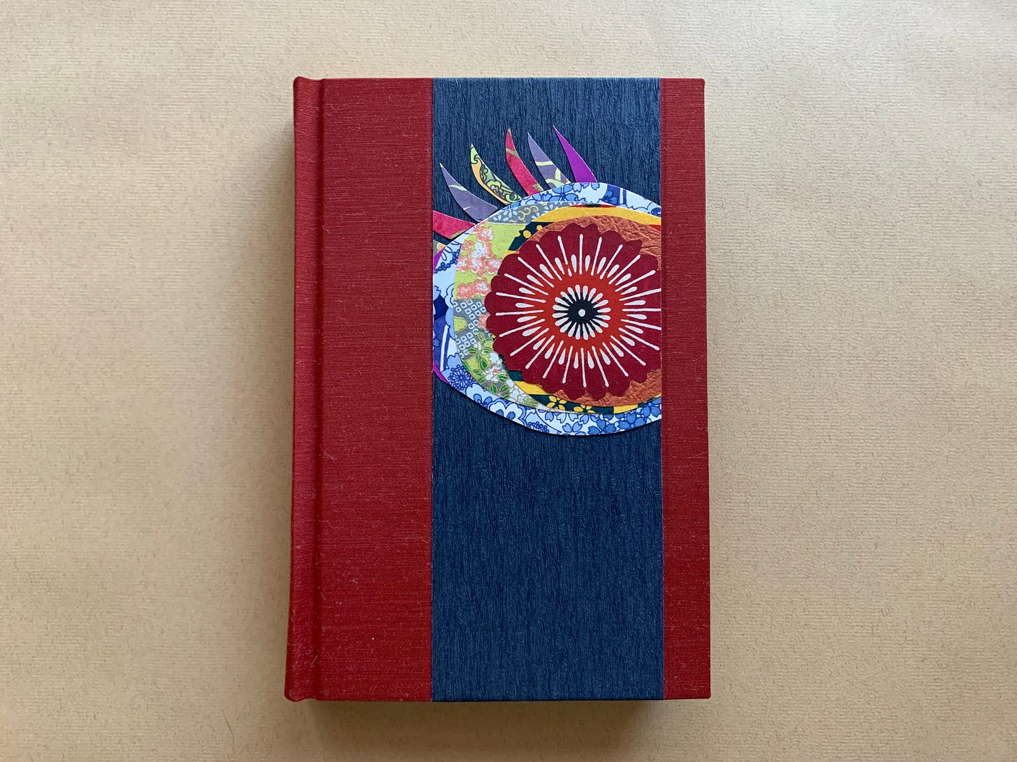 Photo of a handbound copy of ENDPAPERS. The cover features two vertical panels of brick-red silk book cloth with a strip of dark blue paper running between them to the right of center. On the paper is a collage of an eye made from rainbow-colored decorative papers.