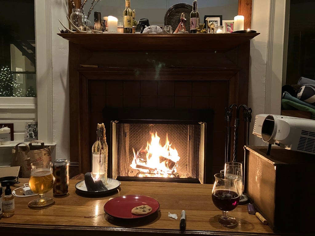 A roaring fireplace and mantle with lit candles. On the coffee table in front of the mantel are two glasses of beer and a plate with a single chocolate chip cookie on it.