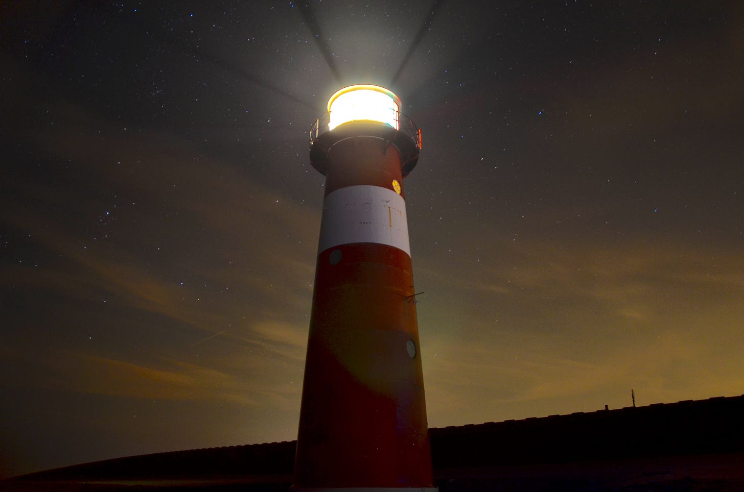 A red and white lighthouse with its light on and shining bright against a darkening sunset sky. Some stars are visible in the sky. A red and white lighthouse with its light on and shining bright against a darkening sunset sky. Some stars are visible in the sky.