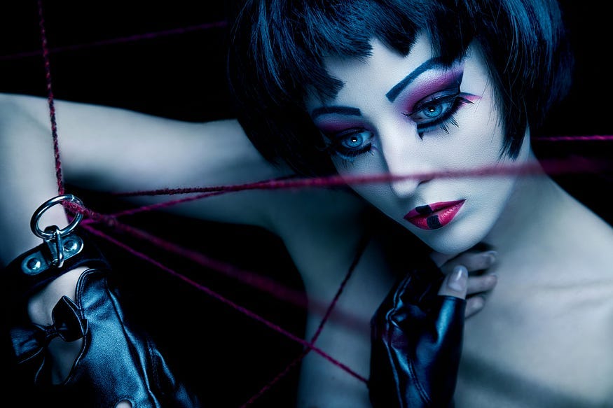 A woman with dark, short hair and doll-like makeup poses artfully, she’s wearing leather gloves, and her right hand is suspended by multiple lines of red rope. The background is black. A woman with dark, short hair and doll-like makeup poses artfully, she’s wearing leather gloves, and her right hand is suspended by multiple lines of red rope. The background is black.