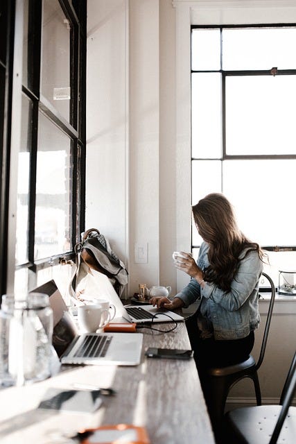Girl sitting at messy desk & typing on laptop, drinking coffee