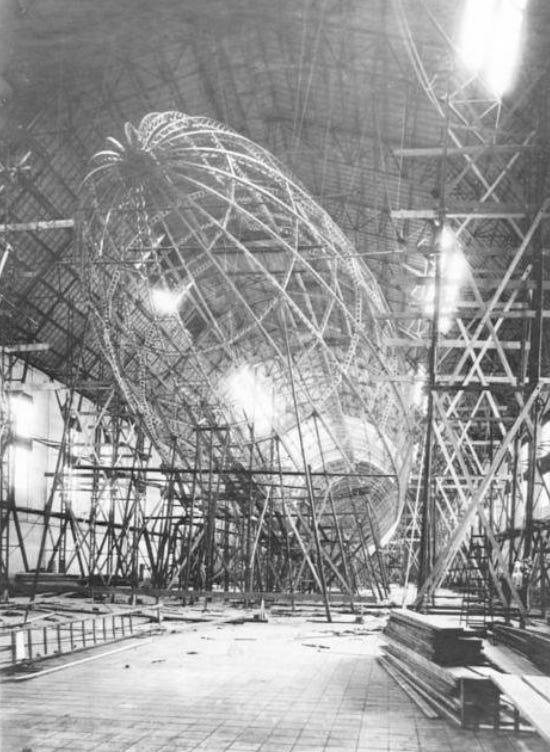 A black and white photo showing a front-end view of the skeletal frame of a Zeppelin whilst under construction in a hangar. The shape of the Zeppelin is clearly visible and is supported by large amounts of wooden scaffolding. 