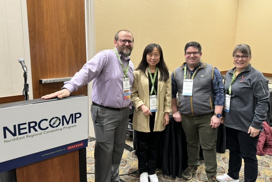 4 People in professional garb stand together in front of a table and to the right of a podium.  They smile at the camera.  From Left to right:  Lance, Xiaorui, Adam, & Stephanie.