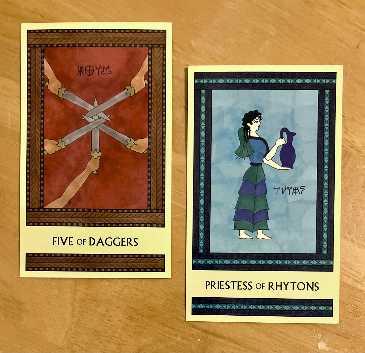 Two Minoan Tarot cards side by side on a golden wood surface. The Five of Daggers is painted in shades of deep red and brown. It shows five hands, each one holding a dagger, with the dagger points woven together in the center of the image, blocking the way. The Priestess of Rhytons is painted in shades of blue. It shows a Minoan woman standing, facing right, holding a large blue pitcher.