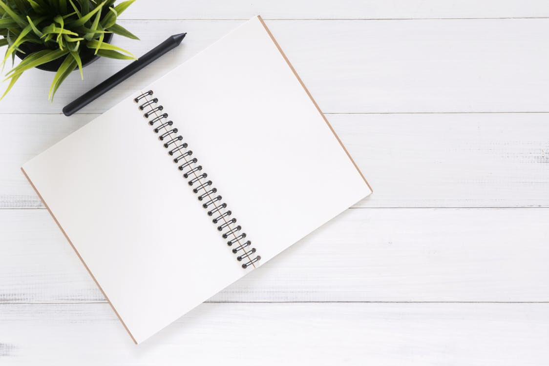 Free Open spiral notebook on white desk with black pen and green plant. Ideal for planning. Stock Photo