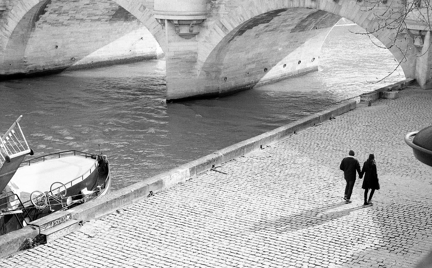 Black and white photograph of a couple, man and woman, walking away from camera below on a cobblestone quay by a river. A boat with a bicycle lashed to it peaks in from bottom left. Two arches of a stone bridge cross the river ahead of the couple and some bare trees branches come into from frame top right