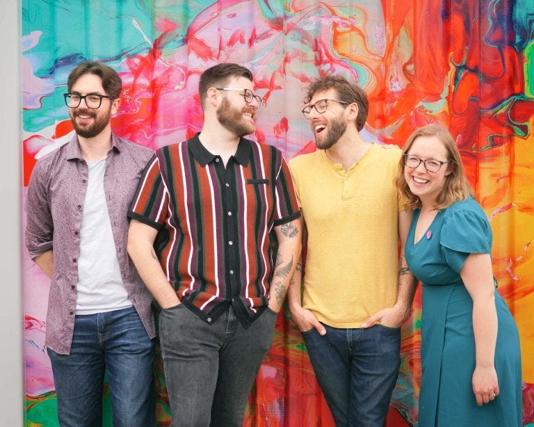 Four people standing and smiling. Background is a colourful mural.