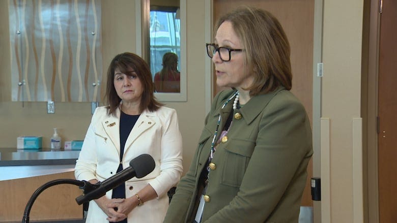 A woman in a green jacket stands in the foreground while a woman with a white blazer stands in the background.