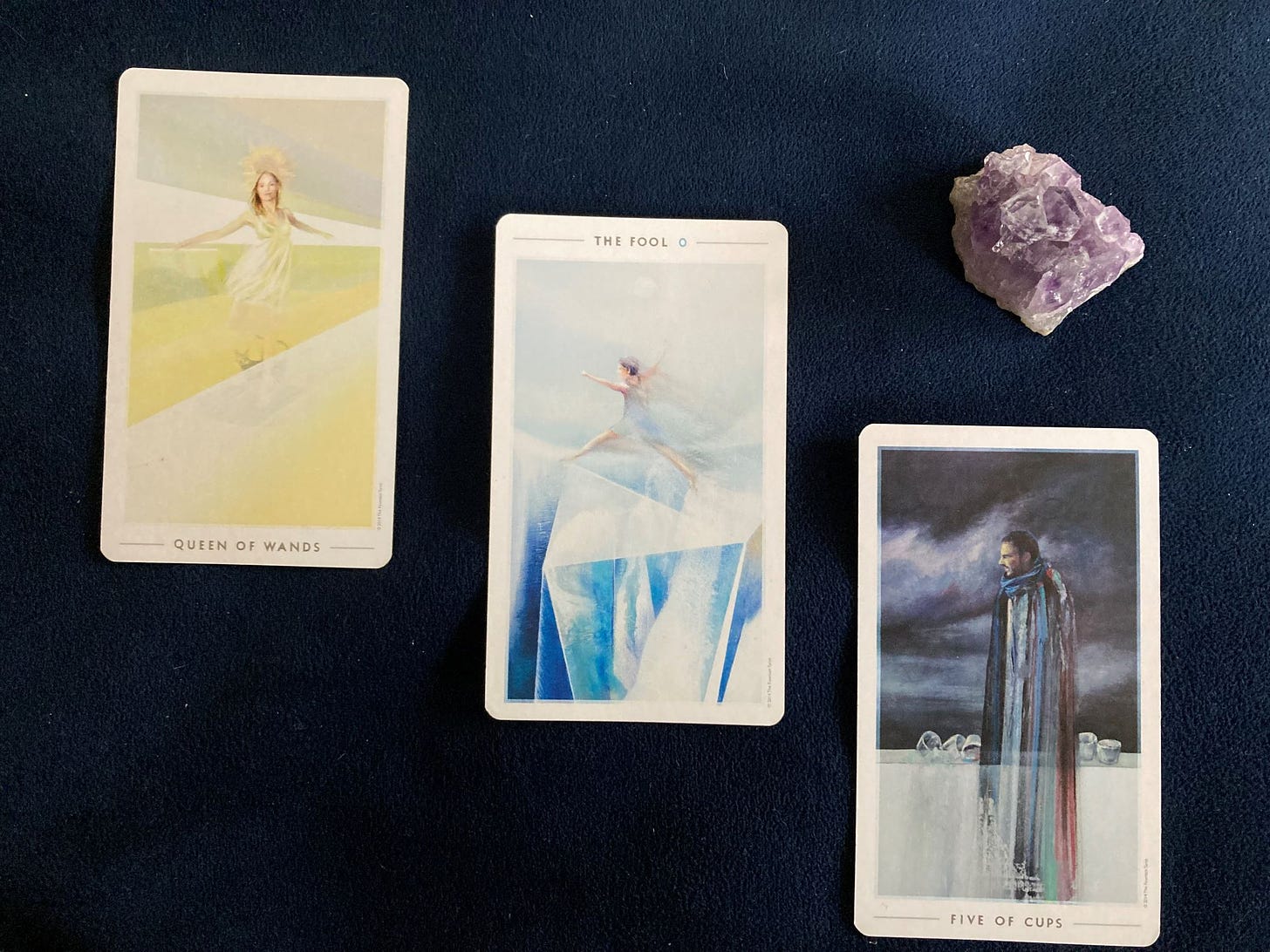 Three tarot cards lay on a blue velvet background with a purple crystal. The cards left to right are Queen of Wands, The Fool and Five of Cups