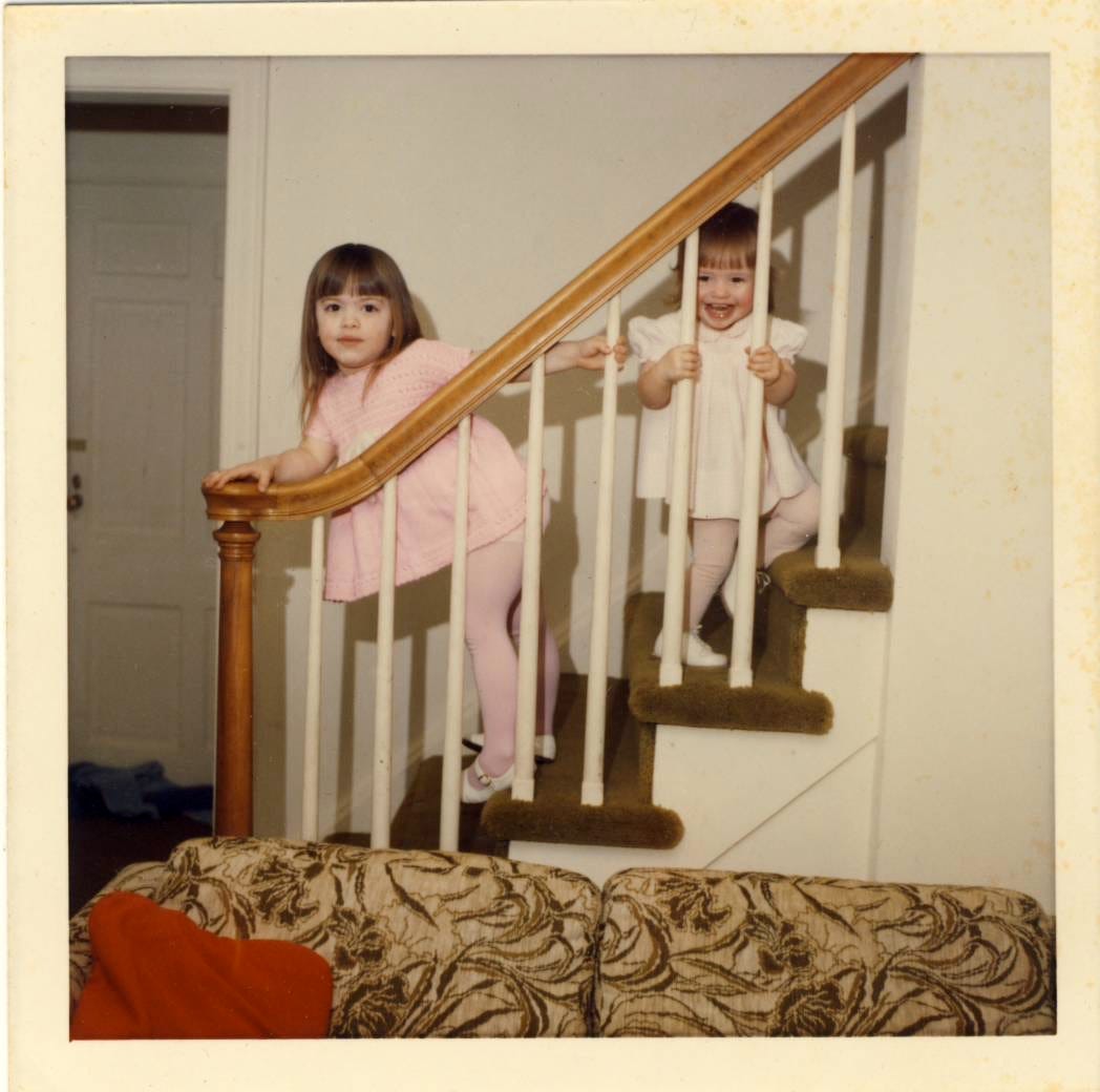Two little girls standing on a staircase, the younger one above, peeking through the railing.