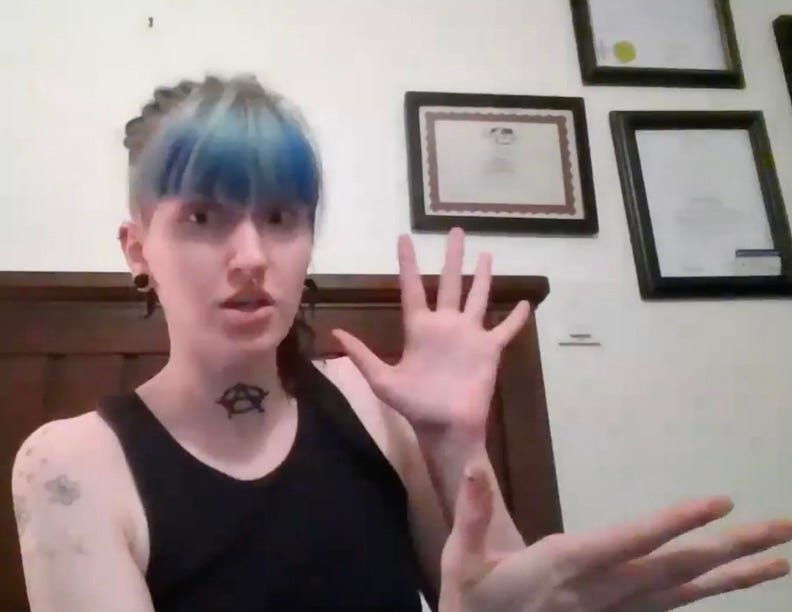 Anna, a young woman with blue ponytailed hair and bangs, gestures against a backdrop of a desk or bed frame. Some awards are visible on the wall behind her to her left, but I've blurred out their contents. She wears a black tank top. She has the anarchy A tattooed on her neck, and some light grey floral tattoos on her right shoulder. Her left arm is up toward her face, fingers outstretched; her right arm is gesturing horizontally to the left