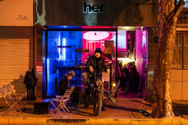 Du Wen sits on a motorbike parked outside a bar at night, with another women sitting on a chair next to her.  