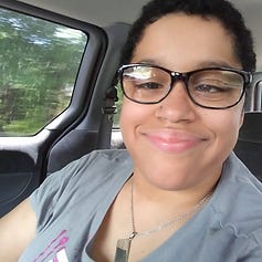 A light-skinned black disabled woman with short hair is wearing black glasses and smiling at the camera from inside a car. It is a sunny day through a window with the green leaves on trees whizzing by.