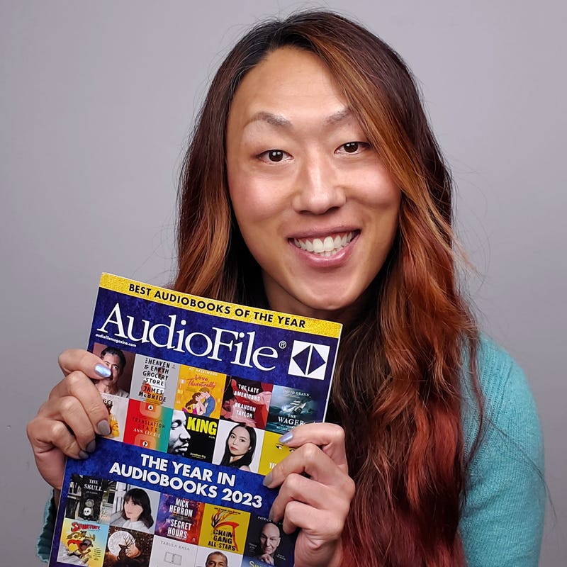 Nicky, smiling, holds the December Issue of AudioFile Magazine, whose cover headlines read: "The Year in Audiobooks 2023" and "Best Audiobooks of the Year”