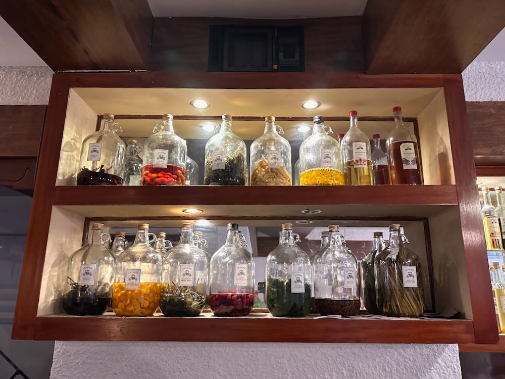 A shelf holding large jugs of a variety of pisco macerados with a variety of colors including red, yellow, and deep green.