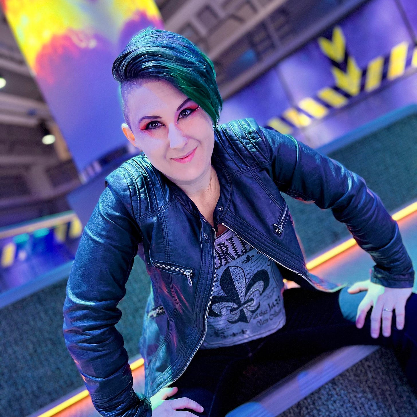 Photo of Lyric Rivera, with green hair and shaved sides, in a black leather jacket, grey shirt, and dark pants, they sit in a purple room with yellow accents that looks like a space ship