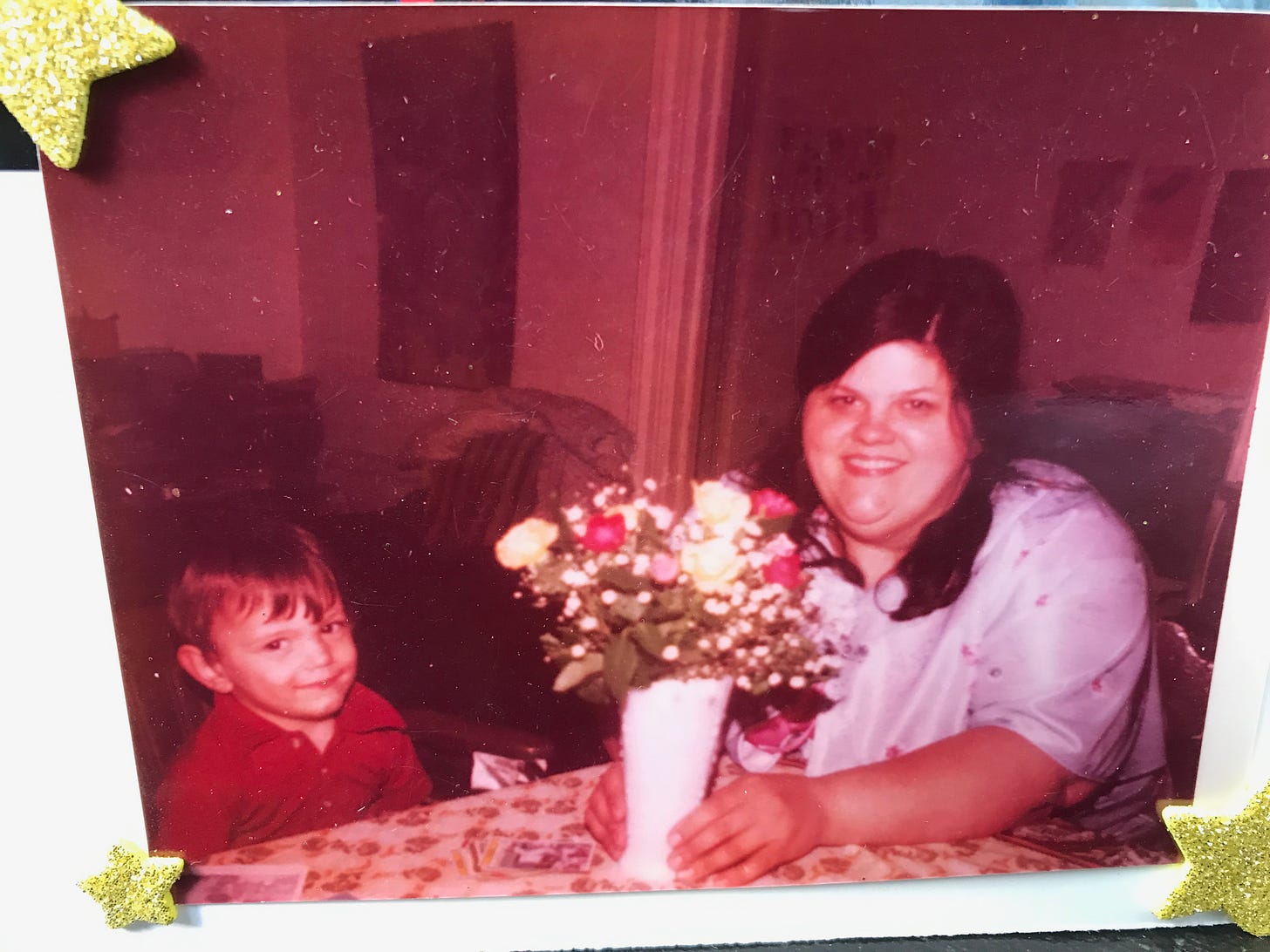 A smiling, dark-haired woman and a smiling, blond little boy sit at a table in a New York apartment. The woman has her hands around the base of a vase holding a bouquet of flowers.