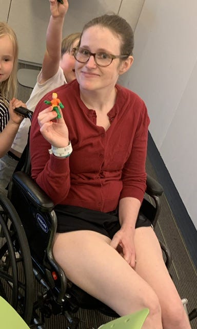 The author, a white woman with brown hair, sits in a wheelchair and holds up a toy with her kids in the background faces cropped out