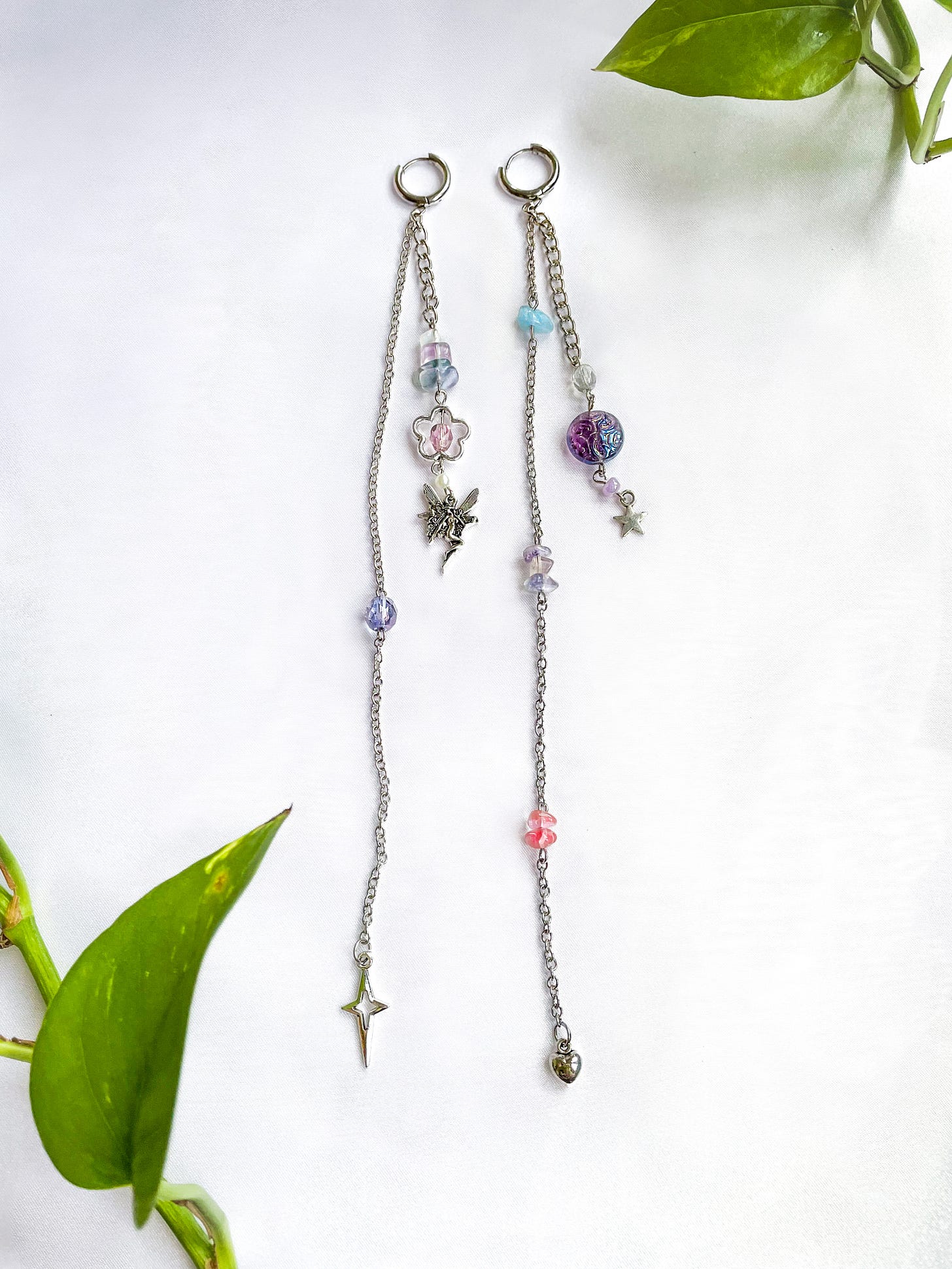 A pair of long mismatched earrings with silver toned jewelry hardware, cable chain, colorful beads and charms lay atop a white cloth background.