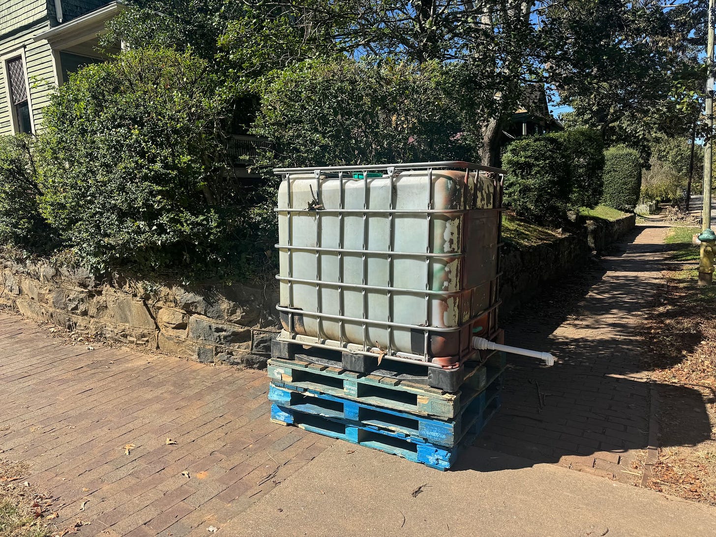 a photo of a large 250-gallon container of water with a spigot propped up on wooden pallets on a street corner with bushes and trees a photo of a large 250-gallon container of water with a spigot propped up on wooden pallets on a street corner with bushes and trees