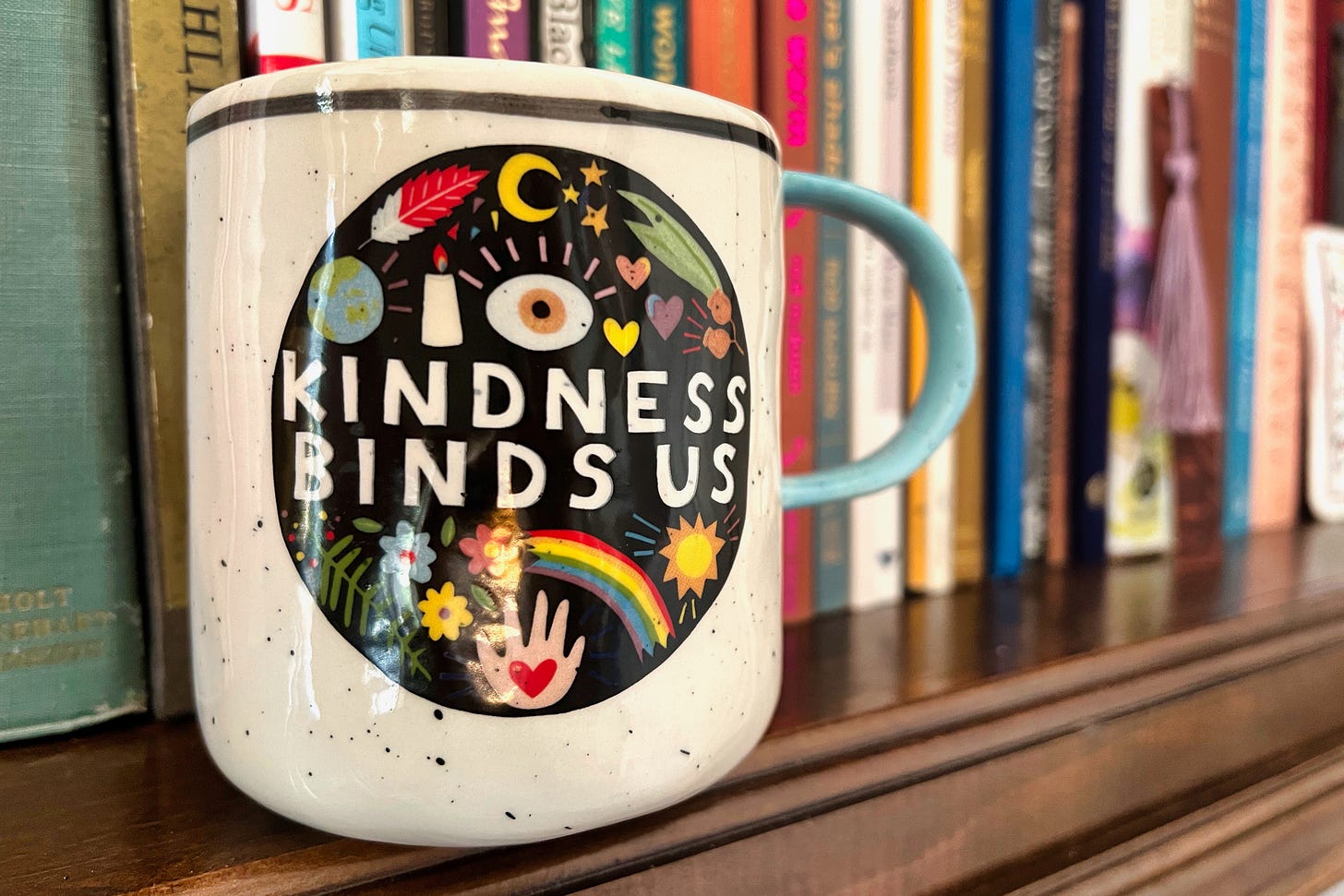 A coffee mug with an oval drawing and these words, “Kindness binds us” sitting on a brown wooden bookshelf with books in the background