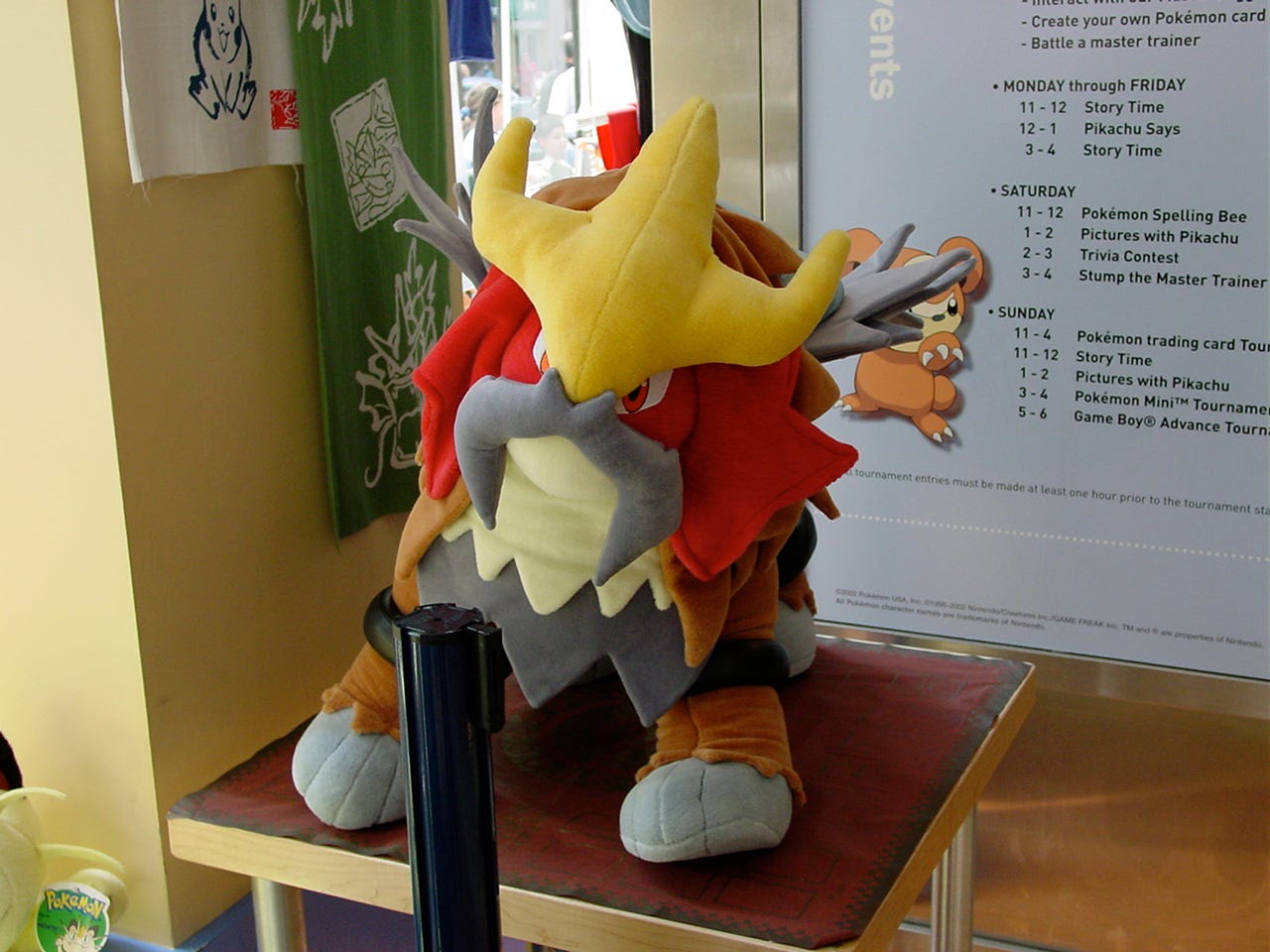 A close-up shot of the huge Entei plush toy that could be seen at the store. On the wall behind it, a schedule with events can be partially seen