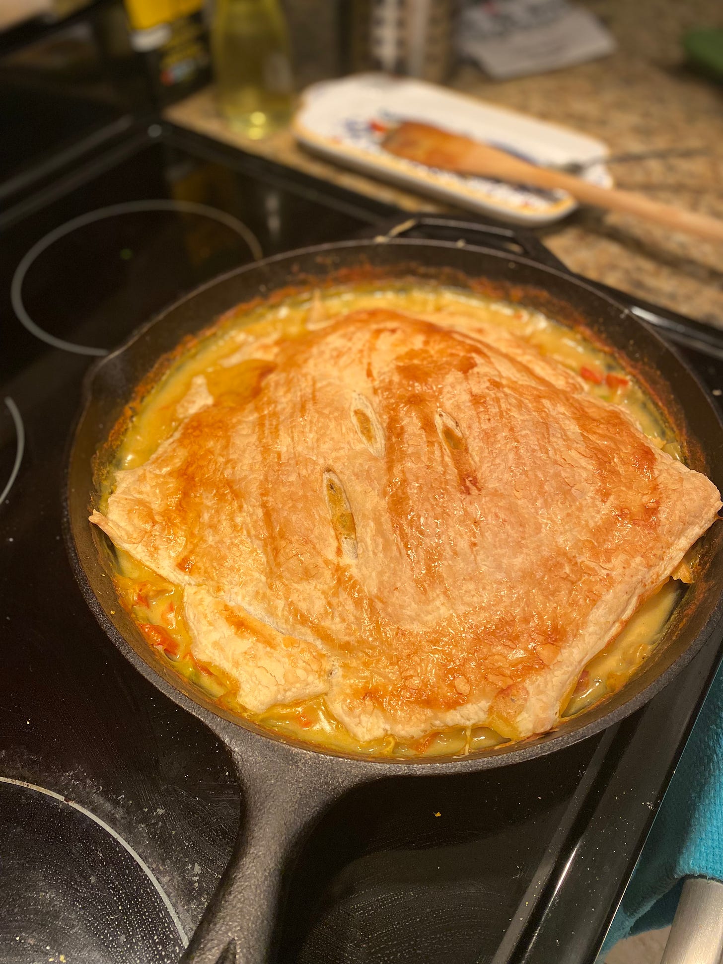 A large cast iron pan with the pot pie described above, a sheet of puff pastry over the top of it with three vents sliced diagonally across the centre. The pastry has shrunk away from the edges of the pan some, so the pie filling can be seen in corners around the pan.