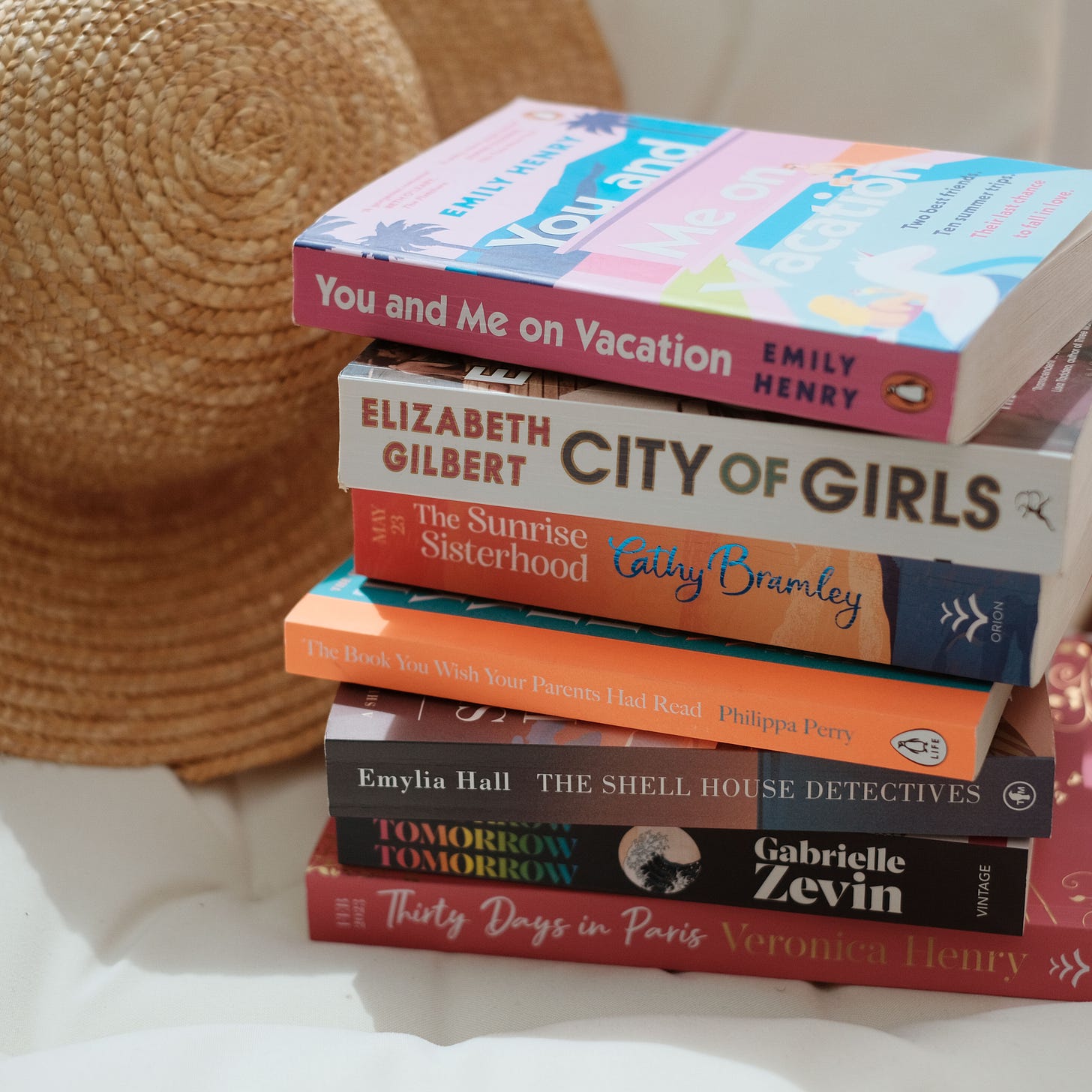 A stack of books on a white cushion. There is a straw hat in the background. The books are You and Me on Vacation by Emily Henry, City of Girls by Elizabeth Gilbert, The Sunrise Sisterhood by Cathy Bramley, The Book You Wish Your Parents Had Read by Philippa Perry, The Shell House Detectives by Emylia Hall, Tomorrow, Tomorrow, Tomorrow by Gabrielle Zevin and Thirty Days in Paris by Veronica Henry.