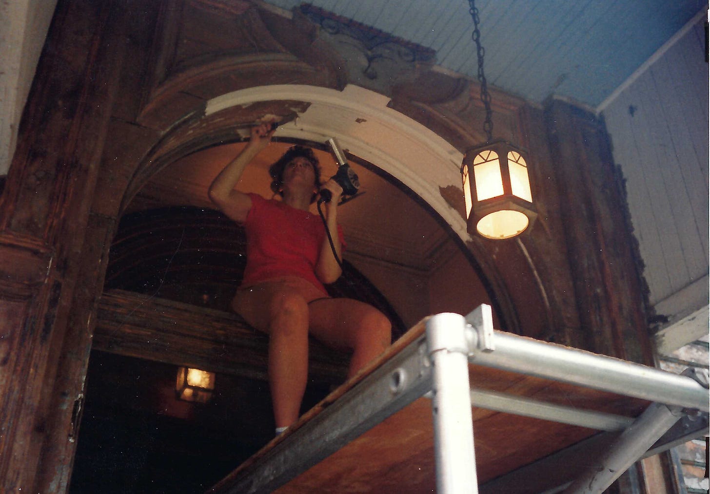 Woman sitting on scaffolding holding a heat gun. She is scraping paint from the archway above her.
