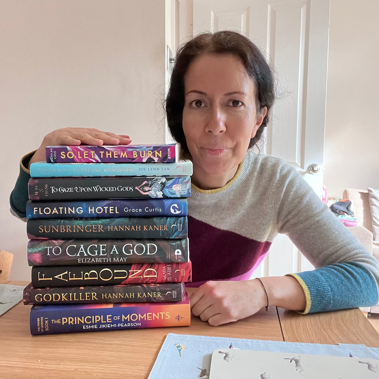 A photo of me next to a stack of books including The Principle of Moments, Godkiller, Faebound, To Cage a God, Sunbringer, Floating Hotel, To Gaze Upon Wicked Gods, Tales of the Celestial Kingdom and So Let The Burn.