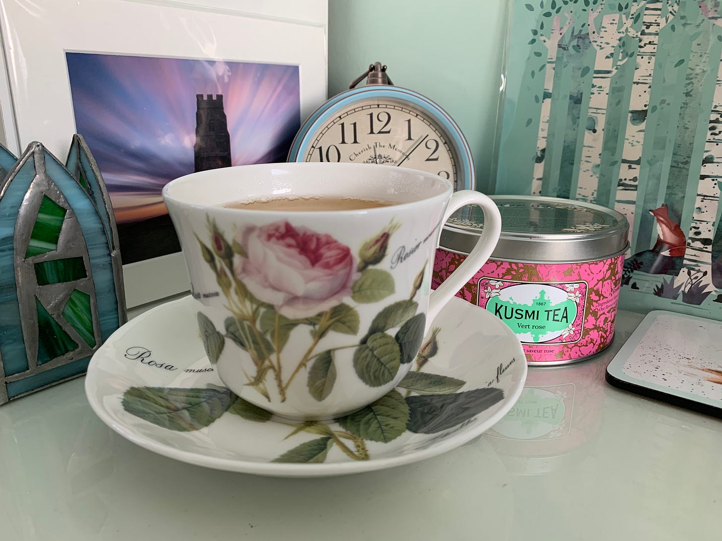 A large bone china tea cup decorated with roses sits on a desk, with a tin of Kusmi Vert Rose tea next to it. The tin is pink and green and looks very posh. Behind them both is a photo of Glastonbury Tor at dawn, a stain glass tea light holder, a clock and a picture of a fox in a wood.