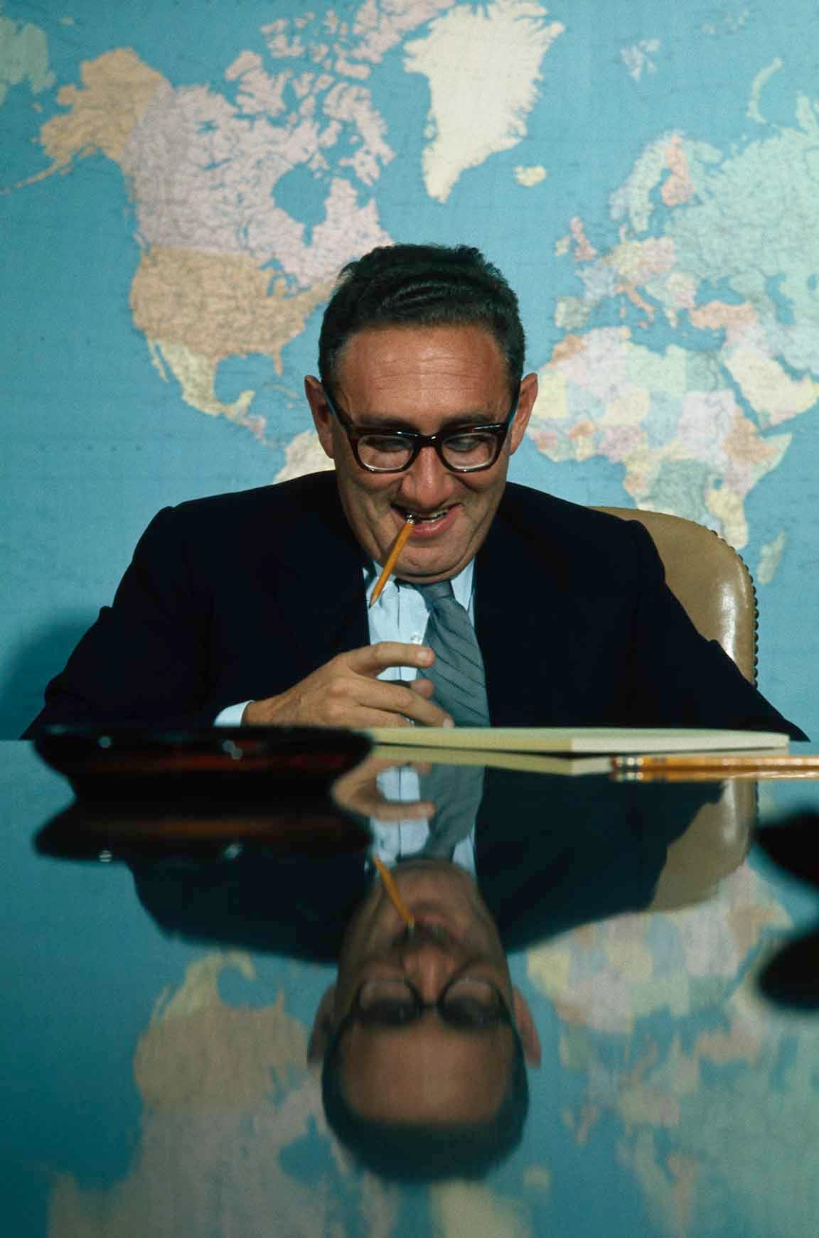 Henry Kissinger circa 1969 sitting at a table with a map of the world behind him. He is looking down, smiling,  with a pencil dangling out of his mouth, and the table reflects his image. White House National Security Affairs Adviser Henry Kissinger poses for a portrait in the Situation Room in the basement of the West Wing at the White House in Washington, D.C. (Photo by © Wally McNamee/CORBIS/Corbis via Getty Images)