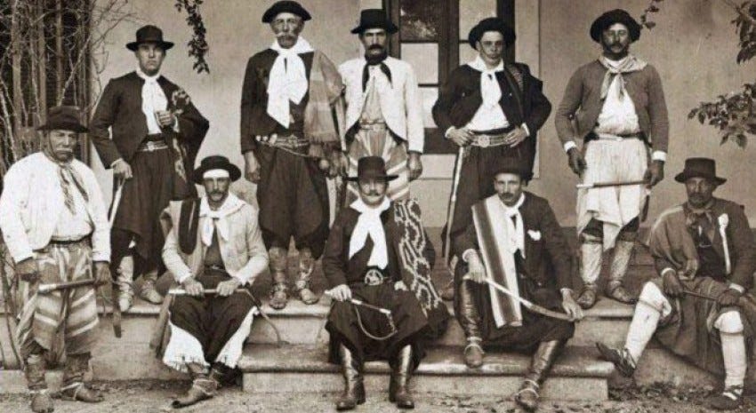 um grupo de homens gaúchos na década de 1920. alguns estão à frente sentados e outros atrás em pé. usam trajes típicos, com bombachas, botas, xales e chapéus. a foto e em preto-e -branco.