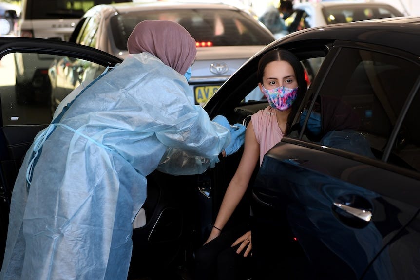 a masked young girl in a car being vaccinated by a nurse