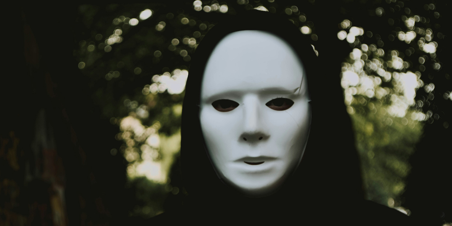 Foto de uma pessoa de máscara branca sem expressão, coberta com um capuz preto, no meio da floresta.