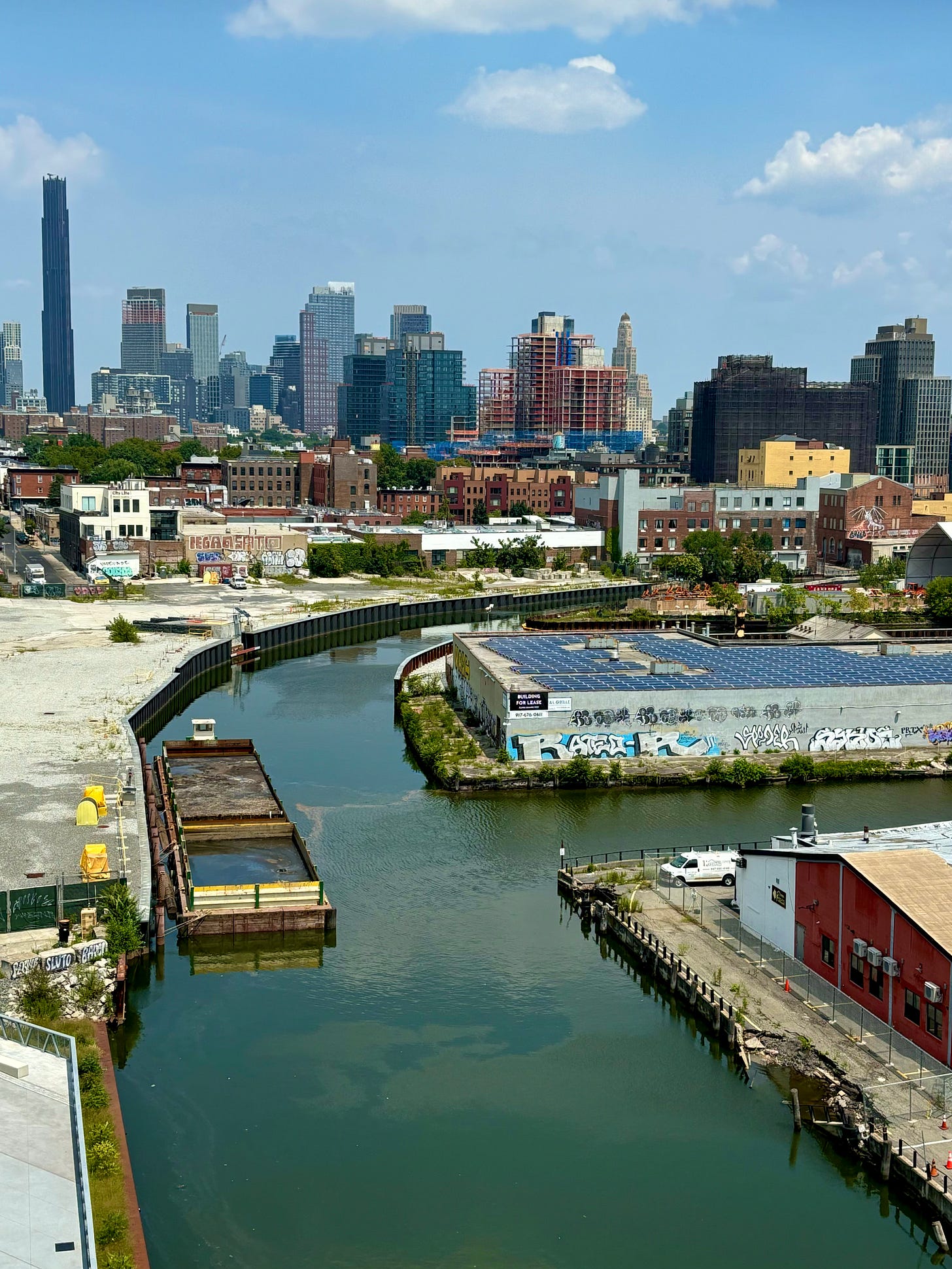 Looking down over Gowanus Canal. Behind it are low houses and further beyond range sky scrapers and many buildings under construction. The sky is blue with some puffy clouds.