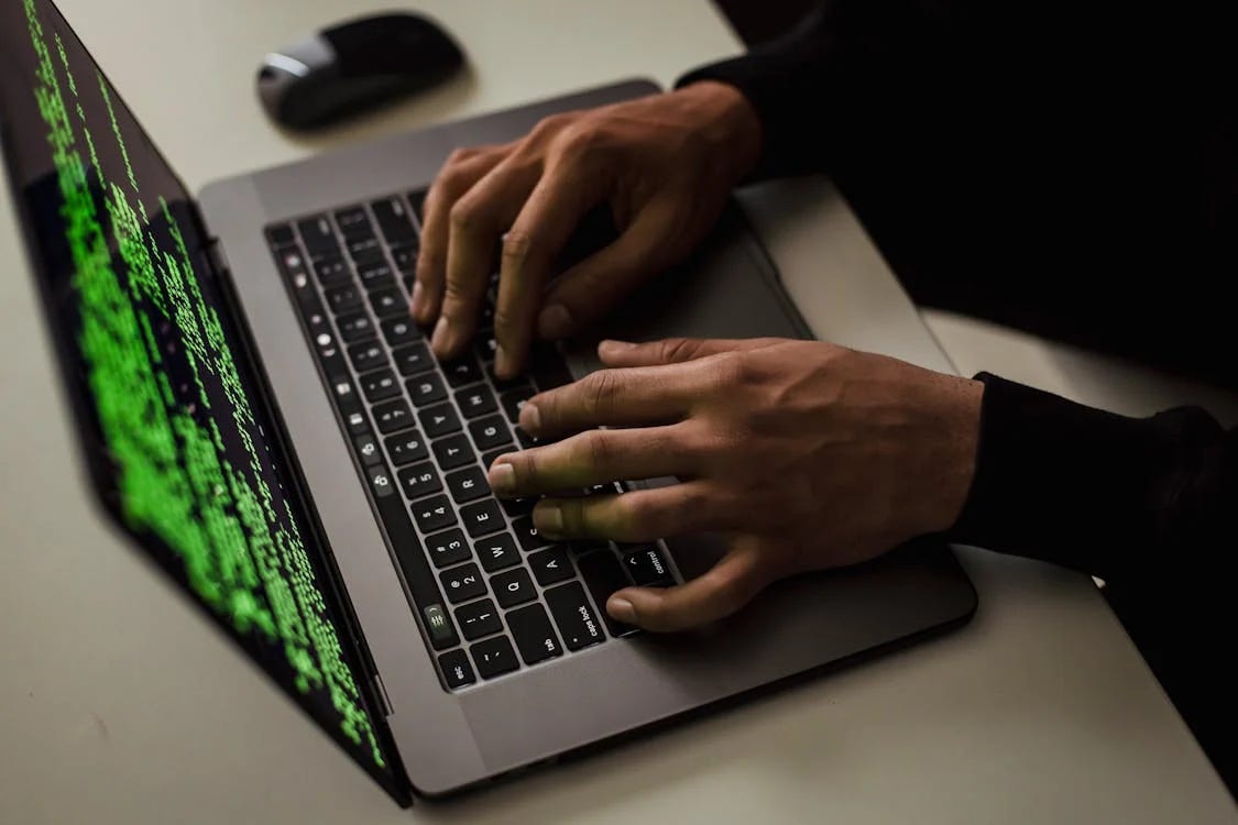 A photo of someone typing on a laptop with a screen of green computer code partially visible.